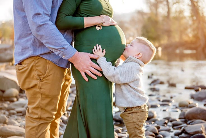 A family by a rocky riverbank, with a man and child touching a pregnant woman's belly.