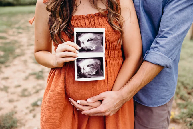 Pregnant woman holds ultrasound images over her belly, man stands behind with hands on her belly.