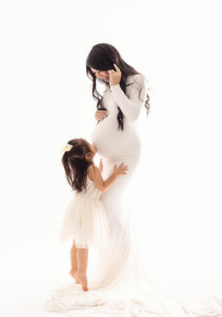 A pregnant woman in a white dress smiles as her young daughter kisses her baby bump.