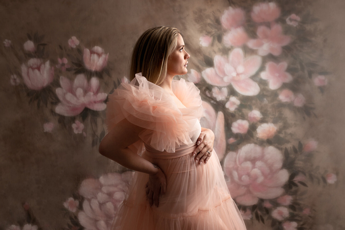 Pregnant woman in a peach tulle gown poses in front of a floral backdrop.