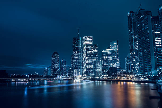 lluminated skyscrapers reflecting on calm water at night, with light trails.