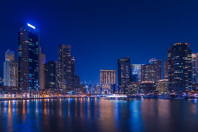 A nighttime cityscape featuring tall, illuminated buildings reflecting on a calm body of water.