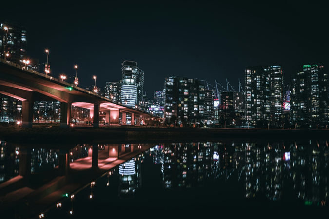 Nighttime city skyline with glowing skyscrapers reflecting on still water.