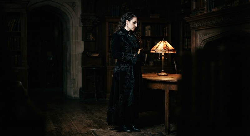 Adult model in a long black gothic coat lit from one side by a warm lamp in a dark room, with deep shadows and a mysterious mood
